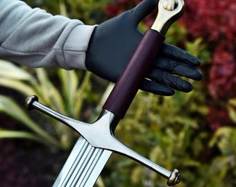 May include: A sword featuring a polished steel blade with a central fuller, a dark maroon leather-wrapped grip, and a brass-toned pommel and crossguard with rounded finials. The crossguard extends outward, and the pommel has a circular design.