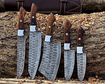 May include: A set of five knives with dark wood handles and silver bolsters, featuring patterned Damascus steel blades. The knives vary in size and shape, including a cleaver, and are displayed with a brown leather roll case in the background.