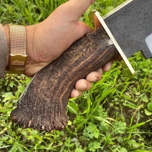 Op de afbeelding: Een vaststaand mes met een donker, getextureerd gewei-handvat en een messing stootplaat. Het lemmet heeft een zwarte, gehamerde afwerking. Het mes wordt vastgehouden door een hand met een goudkleurig mesh horloge met de tekst Lotus op de sluiting.
