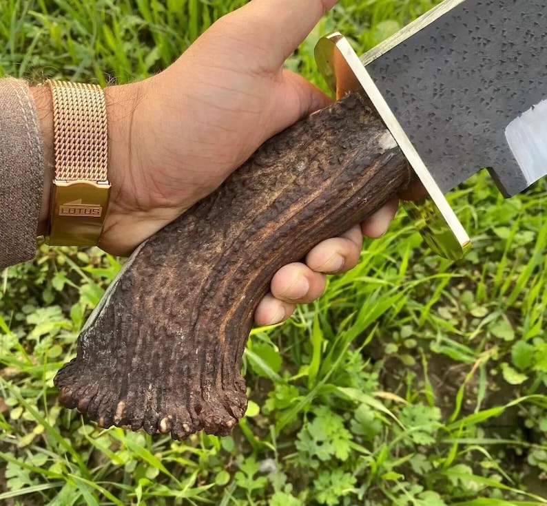 Op de afbeelding: Een vaststaand mes met een donker, getextureerd gewei-handvat en een messing stootplaat. Het lemmet heeft een zwarte, gehamerde afwerking. Het mes wordt vastgehouden door een hand met een goudkleurig mesh horloge met de tekst Lotus op de sluiting.