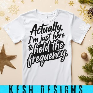 May include: A white short-sleeved t-shirt featuring bold black text printed on the front that reads, Actually, I'm just here to hold the frequency. The shirt has a crew neckline and is made of a smooth fabric with a clean, simple design.