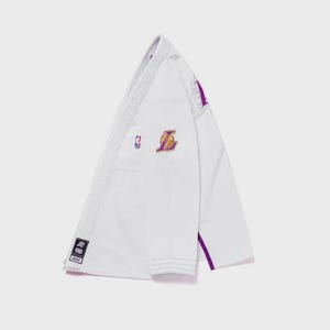 May include: White martial arts uniform with purple and gold accents. Features the Los Angeles Lakers logo and NBA emblem. The uniform has a minimalist design with a purple stripe along the side and a small patch with text.