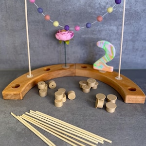 Könnte beinhalten: Ein halbkreisförmiger Holzsockel mit Löchern, zwei vertikale Stäbe mit einer Girlande aus bunten Filzkugeln, eine rosa Blume, eine marmorierte Zahl zwei, mehrere kleine Holzzylinder und eine Sammlung dünner Holzstäbe zum Zusammenbauen.