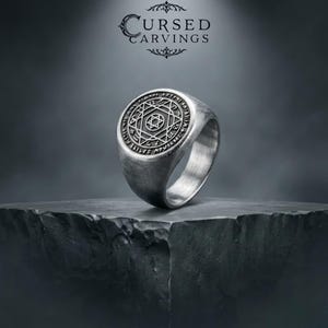 May include: A silver-coloured metal ring featuring a circular face with an engraved six-pointed star enclosed within a geometric pattern and surrounded by a border of small symbols. The band has a brushed finish and a solid, thick construction.