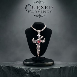 Può includere: Una collana con pendente color argento che presenta un design a spada con ali spiegate e una pietra centrale rossa a forma di cuore. Il pendente pende da una spessa catena a scatola. Sopra il gioiello, il testo CURSED CARVINGS è in nero.