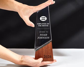 May include: A clear glass employee of the month award with a wooden base. The award has the text "YOUR LOGO", "EMPLOYEE OF THE MONTH", and "AWARDED TO NOAH JOHNSON". The base has the year "2026" engraved on it.