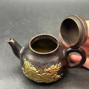 May include: A small, dark-toned metal teapot featuring a rounded body and a curved spout. The exterior is decorated with a raised, gold-colored relief of a dragon. The lid is detached and open, revealing the hollow interior of the vessel.