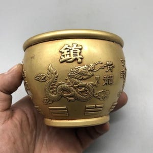May include: A round, gold-colored metal bowl featuring embossed relief designs of a dragon, clouds, and Chinese characters. The exterior surface displays intricate carvings of a mythical creature and geometric patterns around the base.