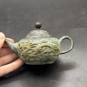 May include: A small, weathered bronze-toned teapot featuring an intricate relief of a dragon winding around its body. The teapot has a rounded lid with a dark knob, a curved spout, and a handle, all displaying a textured, aged metallic finish.