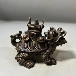 Puede incluir: Una figura de metal color bronce que representa una criatura mítica con un caparazón de tortuga, cabeza de dragón y una estructura en forma de corona en la espalda. La criatura presenta patrones en relieve y dos acentos circulares.
