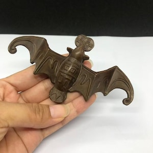 May include: A bronze-coloured metal bat figurine with outstretched wings and a stylised body. The bat features a circular coin-shaped element attached to its head, which is embossed with Chinese characters. The surface has a matte finish.
