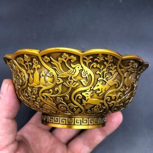 May include: A gold-colored metal bowl featuring an intricate embossed design of birds, deer, and floral motifs. The bowl has a scalloped rim and a base decorated with a geometric Greek key pattern, showcasing a metallic finish and detailed relief.