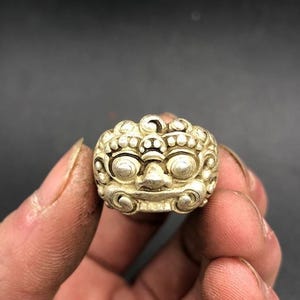 Puede incluir: Un anillo de tono plateado que presenta un rostro esculpido y estilizado con ojos circulares prominentes, una nariz ancha y una melena rizada y texturizada. La superficie metálica tiene un acabado mate con áreas hundidas oscurecidas.