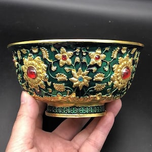 Pode incluir: Uma taça decorativa com acabamento em esmalte verde profundo, apresentando padrões florais e motivos de videira em relevo dourado. O design é acentuado com pedras preciosas circulares vermelhas e azuis, e a base tem um padrão geométrico.