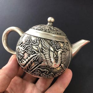 May include: A silver-toned teapot featuring intricate embossed relief carvings of a phoenix bird and stylised clouds on its rounded body. The teapot includes a curved handle, a short spout, and a removable lid with a small spherical knob.