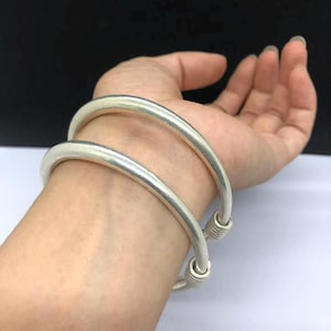 Può includere: Due bracciali rigidi in metallo color argento con una superficie liscia e arrotondata. Ogni bracciale presenta un design semplice e aperto con sottili dettagli a coste alle estremità. I bracciali hanno una finitura metallica lucida.