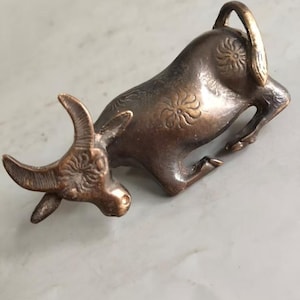 May include: A small bronze-toned metal figurine of a bull in a reclining position. The bull features engraved floral patterns on its head, back, and hindquarters, with a curved tail and textured horns, showcasing a metallic finish throughout.