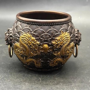 Puede incluir: Un quemador de incienso de metal en tono bronce oscuro con un cuerpo redondeado y un patrón de ondas texturizado. Dos relieves de dragones dorados están centrados en el frente, flanqueados por asas de cabeza de león con anillos de metal.