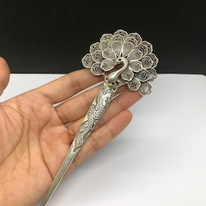 Könnte beinhalten: Eine silberfarbene Haarnadel mit einem detaillierten Pfauenkopf und einem gefächerten Schwanz aus filigraner Drahtarbeit. Der lange, spitz zulaufende Schaft der Nadel ist mit einem geprägten Drachenmotiv verziert, das an der Basis des Pfaus beginnt.