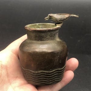 May include: A small, dark bronze-toned metal vessel with a rounded body and a wide opening. The base features a series of horizontal wavy lines embossed into the metal. A small, stylized bird figure is attached to the rim of the vessel.