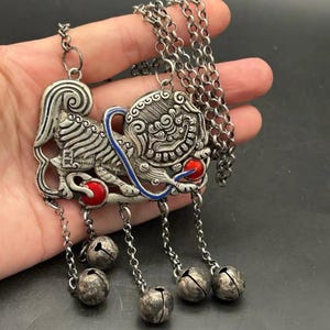 Puede incluir: Un collar con colgante de metal plateado que representa una figura de león guardián estilizado con detalles en esmalte rojo en las patas y una línea curva de esmalte azul. El colgante cuelga de una cadena e incluye cuatro pequeñas campanillas.
