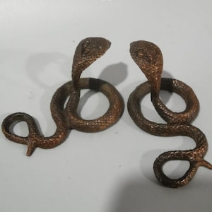 May include: Two bronze-colored metal cobra figurines, each featuring a coiled body and an upright, flared hood. The textured surface of the snakes mimics scales, with detailed markings visible on the heads of both figures in this pair.
