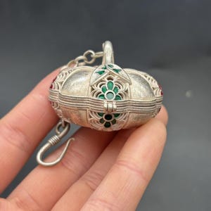 Puede incluir: Un colgante de metal plateado con forma de calabaza, que presenta un intrincado trabajo de filigrana y pequeños detalles en esmalte verde. La pieza incluye un cierre de bisagra, un asa decorativa en la parte superior y un gancho de metal curvo.