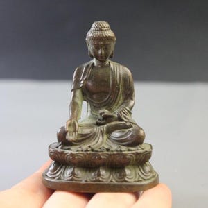 Puede incluir: Una pequeña figura de metal en tono bronce oscuro de un Buda sentado. La figura está representada en una pose meditativa, descansando sobre una base detallada en forma de loto con túnicas texturizadas visibles y expresión serena.
