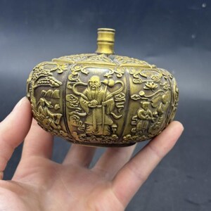 May include: A round, metallic gold-toned vessel with a narrow neck and a flat top. The exterior surface features intricate, raised relief carvings depicting figures in traditional robes, stylised trees, and cloud motifs across its entire body.