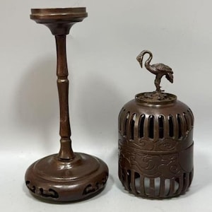 May include: A dark brown metal stand with a tall, slender, bamboo-style stem and a circular base featuring cut-out patterns, paired with a matching cylindrical metal incense burner topped with a sculpted crane figure and cloud motifs.