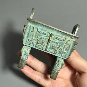 May include: A rectangular bronze vessel with a weathered green patina, featuring intricate raised geometric patterns on its sides and four sturdy legs. The object has two upright handles on the top rim, showcasing a textured, aged metallic finish.