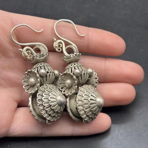 Puede incluir: Un par de pendientes colgantes de tono plateado con intrincado trabajo de metal en filigrana. Cada pendiente consta de un gancho curvo, una pequeña sección decorativa, un elemento central en forma de flor y una base en forma de cúpula.