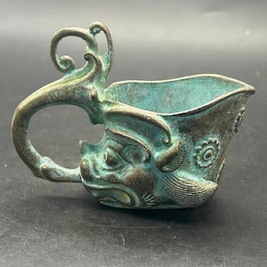Puede incluir: Una copa de metal en tonos bronce con una pátina verde azulada desgastada, que presenta la cabeza de una criatura esculpida en el lateral y un asa grande, ornamentada y curva. El recipiente tiene una parte superior abierta y un motivo floral.