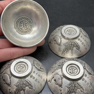 May include: A set of four silver-toned metal bowls featuring embossed fish and plant motifs on the exterior. The base of each bowl displays a circular stamp with a Chinese character, while the interior of one bowl shows a central character.