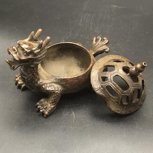 Puede incluir: Un quemador de incienso de metal en tono bronce oscuro con forma de criatura mítica de dragón y tortuga. El cuerpo en forma de cuenco abierto presenta escamas texturizadas, mientras que la tapa extraíble muestra un patrón geométrico.