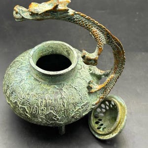 Könnte beinhalten: Ein bronzefarbener Metall-Räuchergefäß mit verwitterter grüner Patina. Das Gefäß hat einen runden Körper, der mit chinesischen Schriftzeichen geprägt ist, und einen drachenförmigen Griff. Ein separater, runder Deckel mit Lochmuster liegt daneben.
