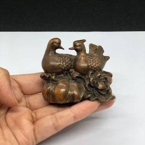 May include: A small bronze-toned figurine depicting two ducks perched atop a rounded base with a sculpted floral element. The ducks feature detailed feather textures and are positioned side-by-side, creating a compact, solid metal ornament.