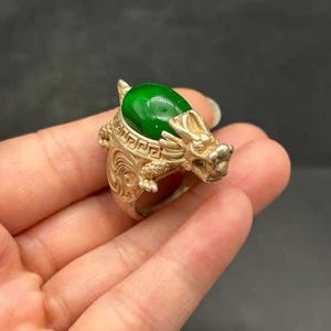 Puede incluir: Un anillo de metal plateado con una piedra ovalada lisa de color verde esmeralda engastada en el centro. La banda está detallada con una cabeza de dragón esculpida en un lado y un patrón geométrico de greca a lo largo del borde superior.