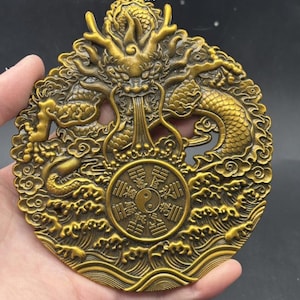 Op de afbeelding: Een ronde goudkleurige metalen plaquette met een gedetailleerd reliëf van een draak omringd door gestileerde wolken en golven. In het midden toont een rond embleem een yin-yang symbool omringd door acht trigrammen met lijnpatronen.