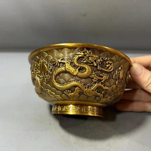Puede incluir: Un cuenco de metal en tono dorado con un intrincado diseño de dragón en relieve que envuelve el exterior. La superficie está decorada con un patrón de ondas repetitivo y un borde geométrico alrededor de la base, mostrando una artesanía detallada.