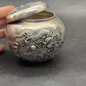 May include: A round silver-toned metal jar with a matching lid, featuring intricate embossed relief designs of a dragon and a phoenix. The surface has a matte finish with detailed carvings of mythical creatures and clouds wrapping around the exterior.