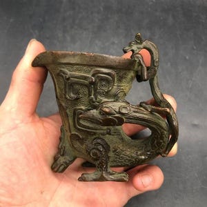 Puede incluir: Un recipiente de metal color bronce con intrincados relieves de patrones en espiral y un asa prominente con forma de dragón. La copa tiene un borde ancho y acampanado, y se apoya sobre una base con forma de criatura mítica con escamas texturizadas.
