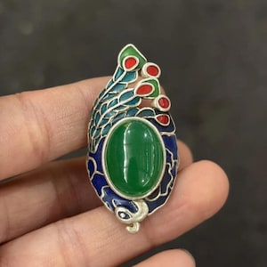 Könnte beinhalten: Eine silberfarbene Brosche mit einem zentralen ovalen grünen Stein, umgeben von blauen, türkisfarbenen und roten Emaille-Akzenten. Das Design umfasst geschwungene, blattartige Muster und kleine kreisförmige Motive, die ein detailliertes Schmuckstück bilden.