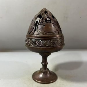 May include: A dark bronze-toned metal incense burner featuring a dome-shaped lid with intricate cut-out patterns and swirling engravings. The base is a pedestal stand with a circular foot, and the body is decorated with a repeating scroll motif.