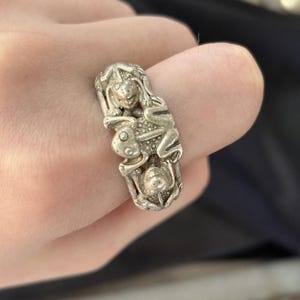 Puede incluir: Un anillo de metal plateado con un diseño detallado en tres dimensiones de tres pequeñas figuras de ranas estilizadas apiladas verticalmente. La superficie del anillo tiene un acabado mate texturizado con detalles en relieve visibles.