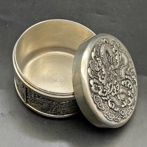 Può includere: Un contenitore rotondo in metallo color argento con coperchio rimovibile. Il coperchio presenta un design a drago in rilievo circondato da nuvole. Anche il lato del contenitore mostra un motivo a drago in rilievo. L'interno è liscio e metallico.