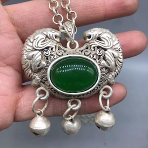 Op de afbeelding: Een zilverkleurige metalen hanger met een centrale ovale groene steen. Het metalen frame is versierd met twee symmetrische tijgerfiguren en ingewikkeld krulwerk. Drie kleine zilverkleurige bolvormige belletjes hangen onderaan de hanger.