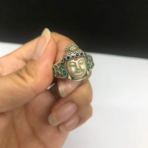 Op de afbeelding: Een zilverkleurige ring met een gebeeldhouwd Boeddha-hoofd als middelpunt. Het ontwerp bevat een rij kleine blauwe ronde accenten langs de haarlijn, een rode stip op het voorhoofd en turquoise emaille patronen aan de zijkanten van de ring.