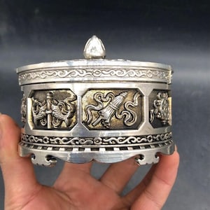 Könnte beinhalten: Ein runder, silberfarbener Metallbehälter mit einem Deckel, der eine spitze Verzierung aufweist. Die Außenseite zeigt eine Reihe achteckiger Felder mit erhabenen Reliefs von Fischen, einem Regenschirm und anderen Symbolen auf dunklem Grund.