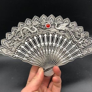 May include: A silver-toned metal hand fan featuring intricate embossed designs of a dragon and a phoenix amidst clouds. The fan has a central circular red gemstone accent and a series of vertical, pointed slats radiating from the base.